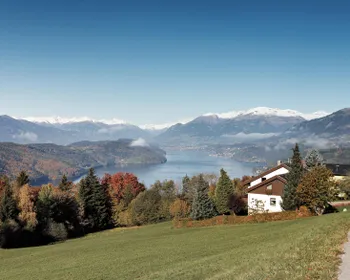 Ein Haus steht halb verborgen in einem herbstlichen Wald, vor einem See und Bergen