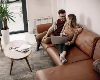 Ein Pärchen sitzt zusammen auf einer Couch. Die Frau hat den Laptop am Schoß und sie sprechen miteinander.