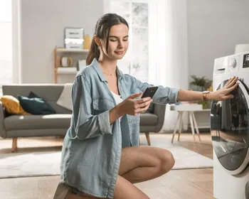 Eine Frau kniet neben der Waschmaschine, hat ihr Smartphone in der Hand und schaut darauf.