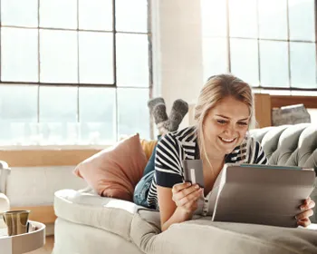Eine Frau liegt auf der Couch und hat ein Tablet und eine Kreditkarte in der Hand.