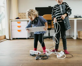 Zwei Kinder sind in einem Büro und haben je eine Tastatur in der Hand. Sie stehen in Mitten von Netzverteilern und Kabeln. Die Kinder sind gut gelaunt.