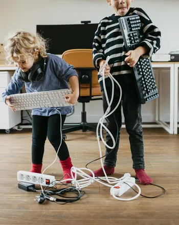 Zwei Kinder sind in einem Büro und haben je eine Tastatur in der Hand. Sie stehen in Mitten von Netzverteilern und Kabeln. Die Kinder sind gut gelaunt.