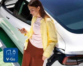 Eine Frau in einem gelben Shirt lehnt an einem weißen Elektroauto und schaut dabei auf ihr Handy. Das Auto wird gerade aufgeladen, und ein V-Go-Logo ist zu sehen.