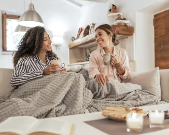 Zwei Frauen sitzen in eine Decke gekuschelt mit je einer Tasse auf der Couch. Vor ihnen steht ein Tisch mit Kerzen und Keksen.