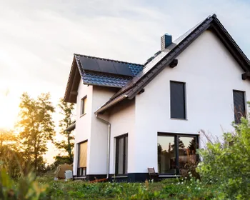 Ein Haus mit einem schwarzem Dach und einer PV-Anlage drauf. Im Hintergrund scheint die Sonne.