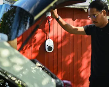 Links im Bild ist eine junge Frau, die neben ihrem E-Auto geht. Rechts sieht man eine Wallbox an einer Holzfassade eines Hauses montiert.