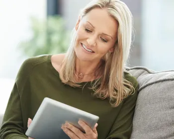 Eine Frau sitzt mit einem Tablet in der Hand auf einer Couch und liest am Tablet.
