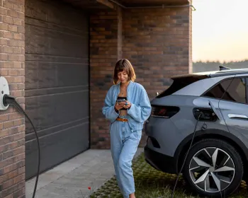 Eine junge Frau steht neben einem E-Auto, lädt dieses und sieht währenddessen auf ihr Smartphone.