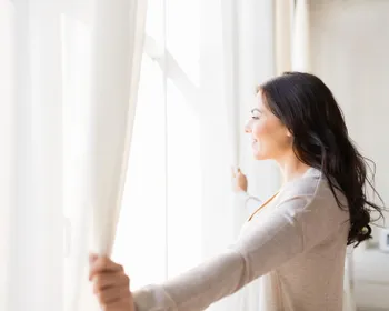 Frau steht vor einem Fenster und öffnet die Vorhänge.