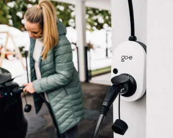 Eine go-e Wallbox ist an einer Hausmauer befestigt. Unscharf im Hintergrund ist eine junge Frau die ein E-Auto ladet.