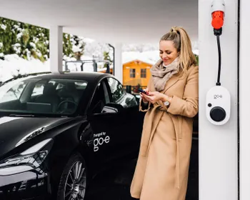 Frau lehnt an einer Hausmauer und sieht auf ihr Smartphone. An der Hausmauer ist eine Wallbox befestigt. Daneben steht ein E-Auto.