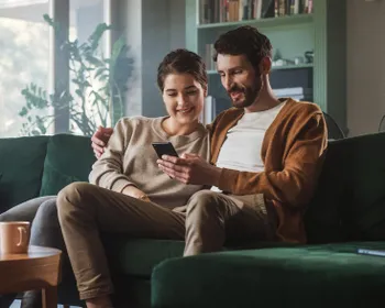 Junges Paar sitzt am Sofa und schaut gemeinsam aufs Smartphone.