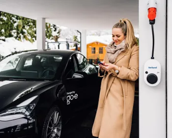 Frau lehnt an einer Hausmauer und sieht auf ihr Smartphone. An der Hausmauer ist eine Wallbox befestigt. Daneben steht ein E-Auto.