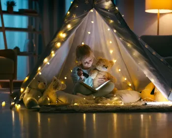 Mädchen sitzt im Dunkeln mit Taschenlampe und Teddy in einem Zelt mit Lichterkette und liest ein Buch.