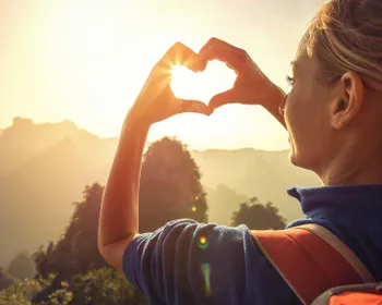 Eine Frau mit Wanderrucksack ist von hinten zu sehen und sie blickt auf den Sonnenuntergang. Mit den Händen formt sie ein Herz, dass die vor die Sonne hält.