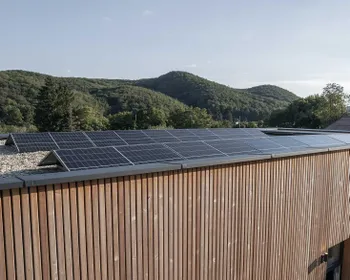 Moderns Haus mit Holzfassade und Flachdach. Am Dach ist eine PV Anlage. Im Hintergrund ist ein Wald.