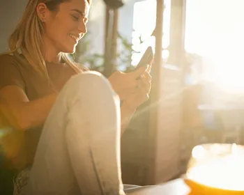 Junge Frau sitzt auf einem Sessel mit Handy in der Hand und die Sonnenstrahlen scheinen bei der Balkontür rein.