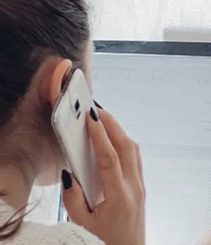 Eine Frau ist von hinten zu sehen. Sie sitzt vor einem Computer und telefoniert mit dem Handy.