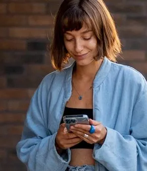 Eine junge Frau steht neben einem E-Auto, lädt dieses und sieht währenddessen auf ihr Smartphone.