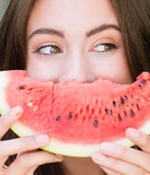 Frau hält ein Stück Wassermelone vor ihrem Gesicht und sieht glücklich aus.