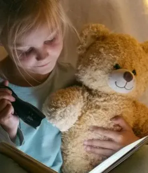 Mädchen sitzt im Dunkeln mit Taschenlampe und Teddy in einem Zelt mit Lichterkette und liest ein Buch.