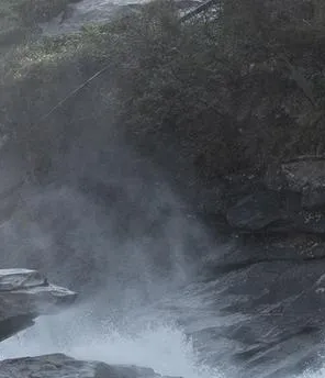 Ein kleiner Wasserfall bei einem Gebirgsfluss. Der Fluss liegt zwischen Wäldern.