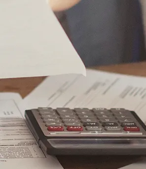 Auf einem Tisch liegen Dokumente, ein Taschenrechner und ein Laptop. Zwei Personen lehnen darüber, eine hält ein Blatt Papier