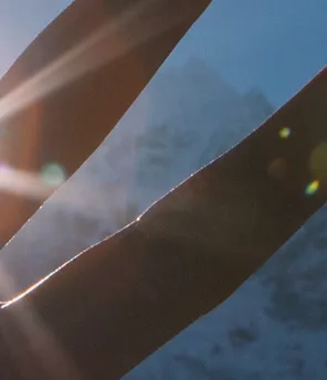 Vor dem Hintergrund schneebedeckter Berge unter strahlend blauem Himmel verdeckt eine Hand teilweise die Sonne, wobei Sonnenstrahlen zu sehen sind.