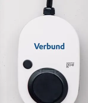 Frau lehnt an einer Hausmauer und sieht auf ihr Smartphone. An der Hausmauer ist eine Wallbox befestigt. Daneben steht ein E-Auto.